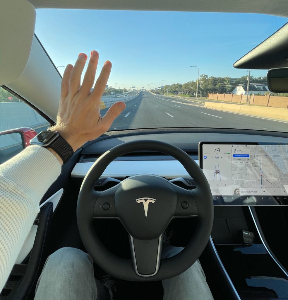 Tesla car driving down Texas toll road, with steering wheel in bottom middle, taken from driver's perspective and hand waving