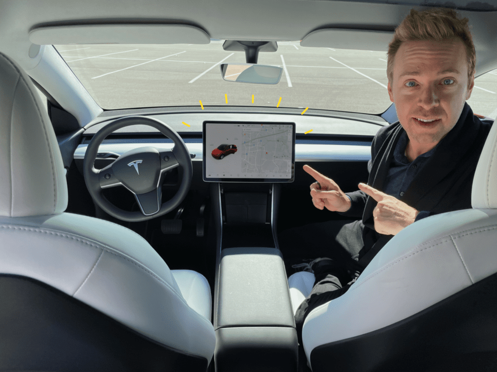 Blonde haired, blue-eyed attractive man sitting in passenger seat of a car, pointing to a center LCD screen (Tesla Model Y center console feature photo)