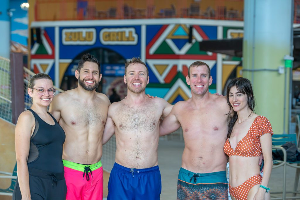 five adults in their 30s (and one in their 40s, me) standing in a group with arms around each other and all in bathing suits