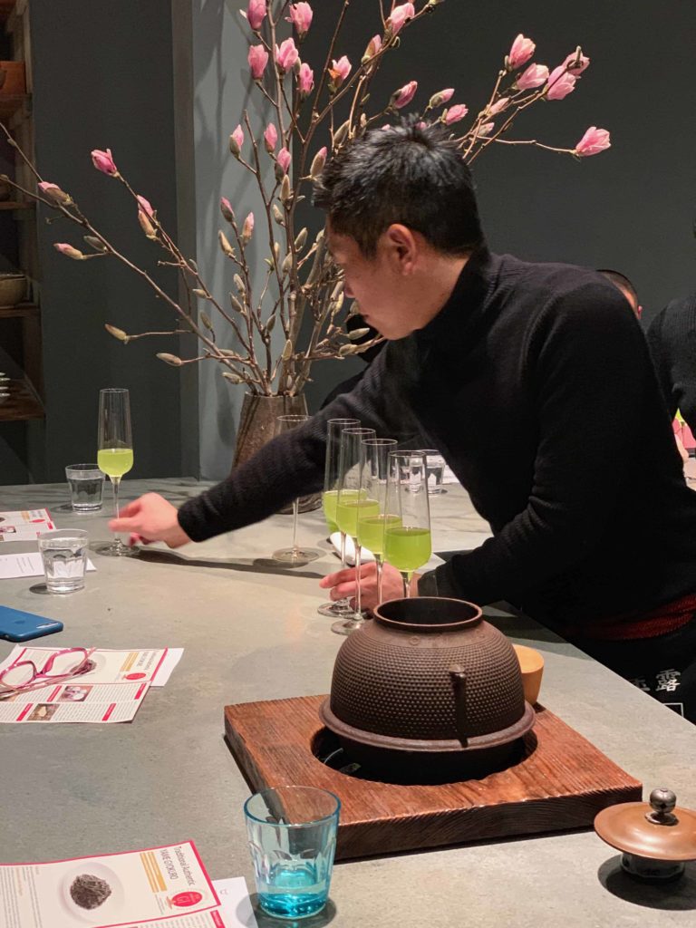 Man in black turtleneck, many long-stemmed champagne glasses, and a pretty pink floral arrangement behind him.