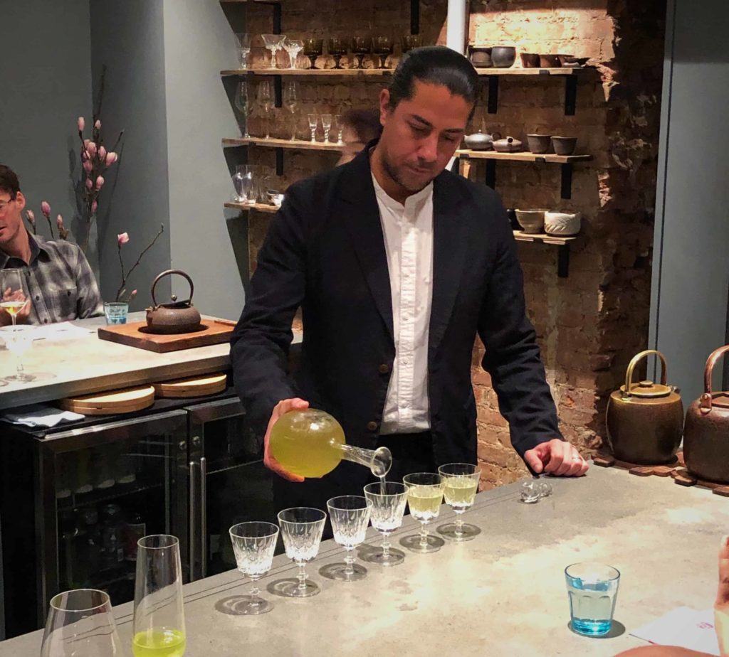 Man wearing a black jacket, pouring green liquid into about seven glasses all lined up