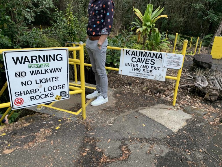 Lower half of a man wearing pants and a shirt, and signs that way WARNING and KAUMANA CAVES