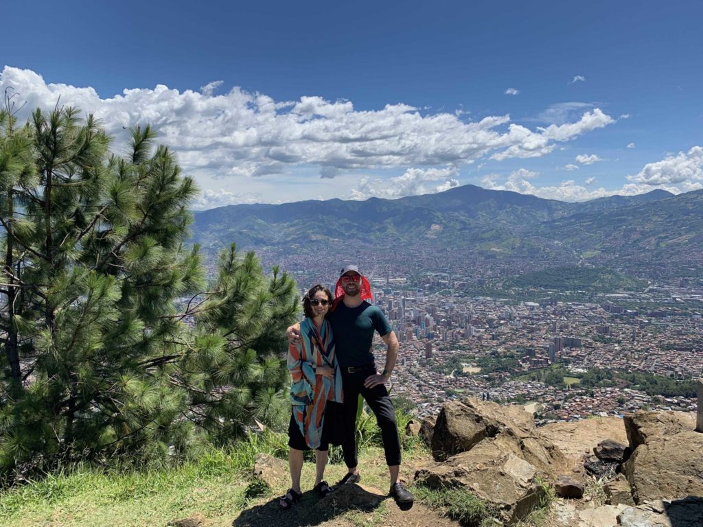 Two people standing at top of hill overlooking city below, clouds and green stuff