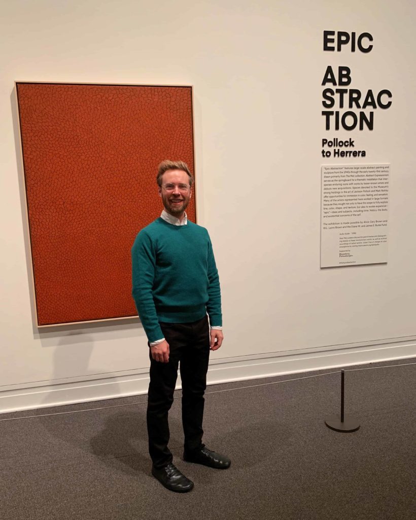 Man in green sweater standing in front of a red painting inside the Met Museum on a white wall