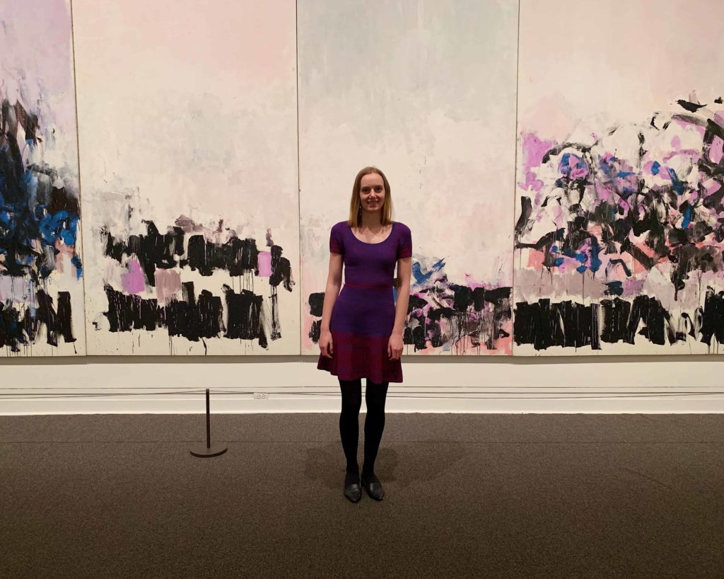 Blonde woman in a purple-ish dress in front of a few panels of a painting