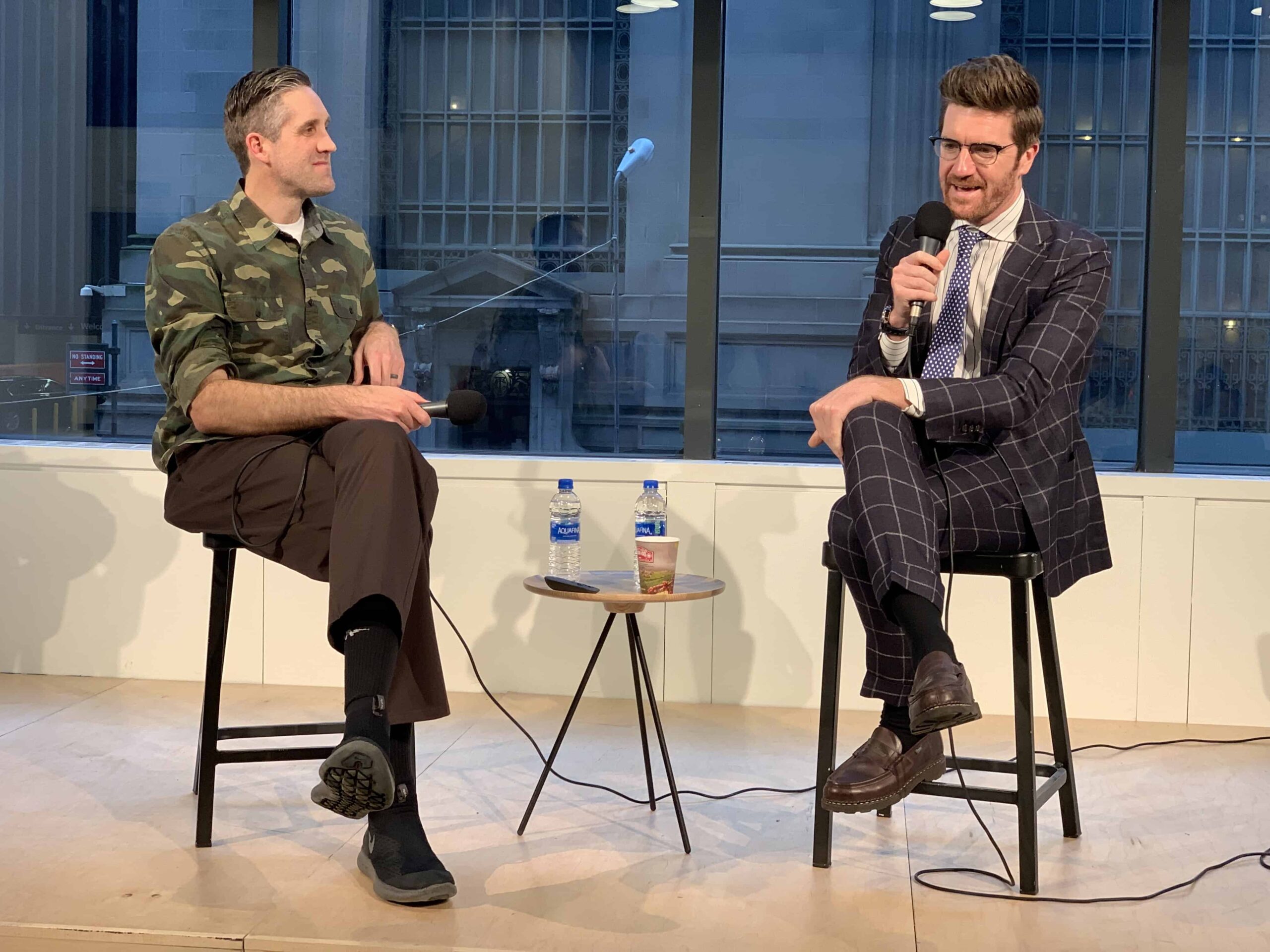 Two tall, dapper men on stage sitting in chairs holding microphones