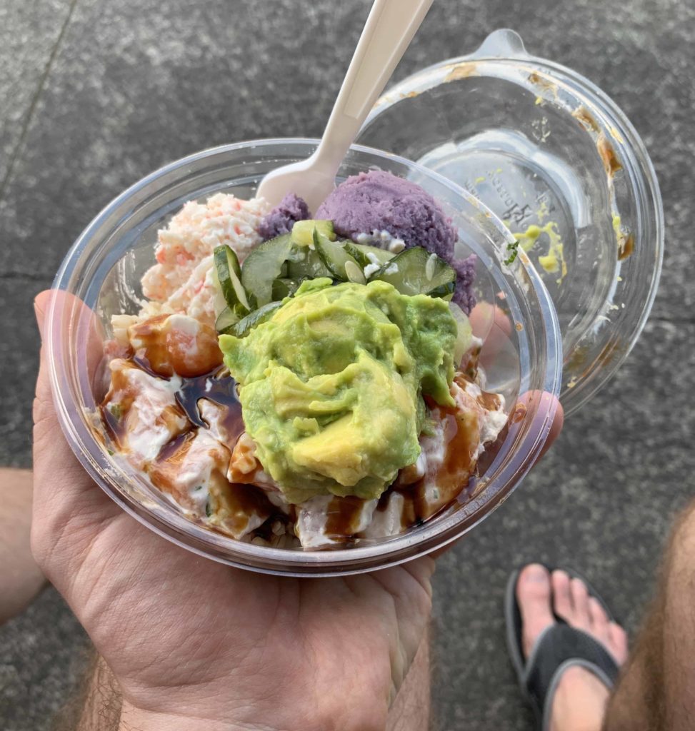 Hand holding a plastic container that has poke and green avocado and purple scoop of potato