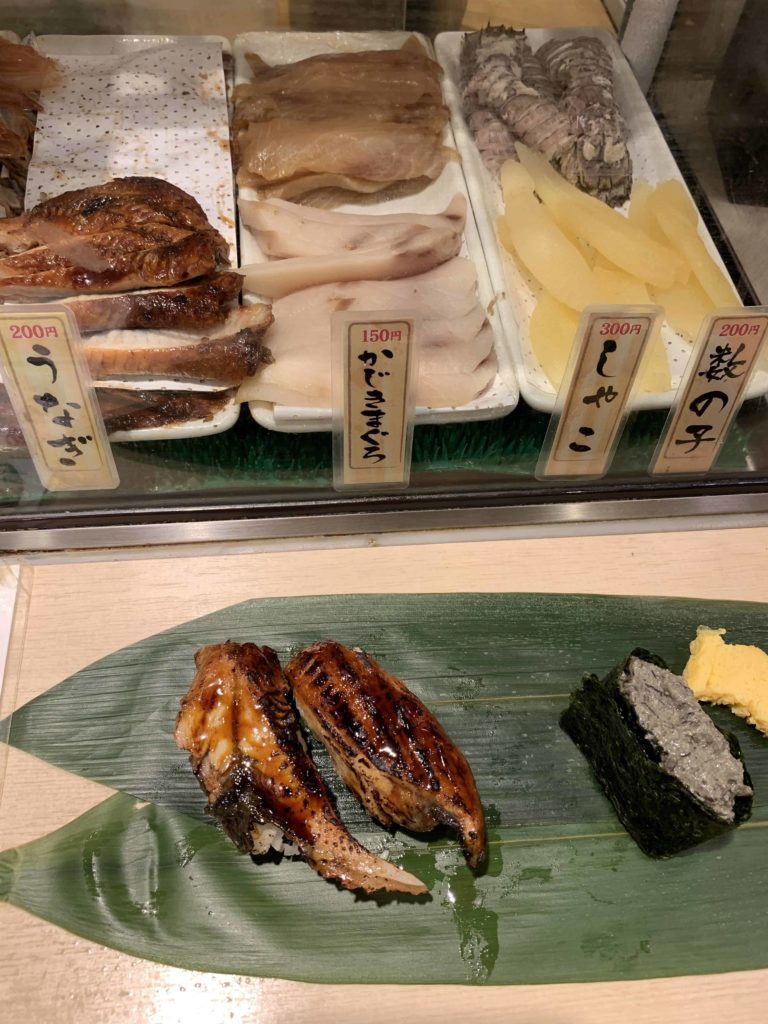 Banana leaf has three pieces of sushi on it, plus case with raw fish inside, pictured at Standing Sushi in Shinjuku