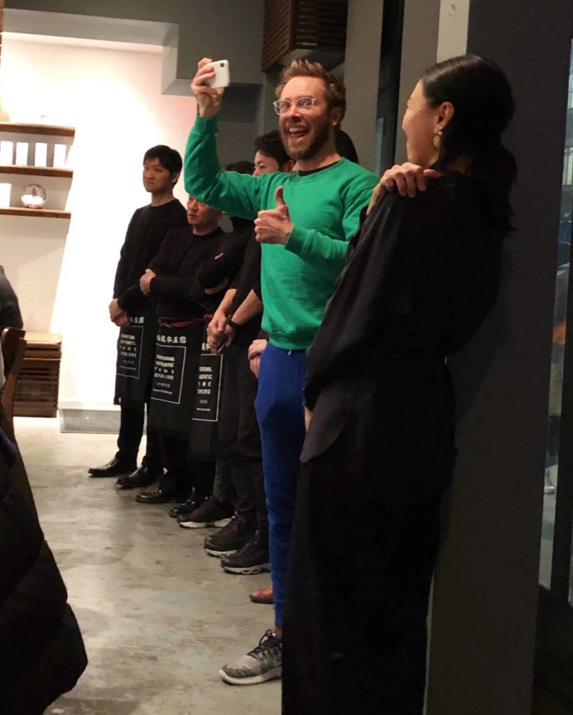White man (me) wearing green sweatshirt, blue sweatpants, surrounded by lots of Japanese staff all wearing black.