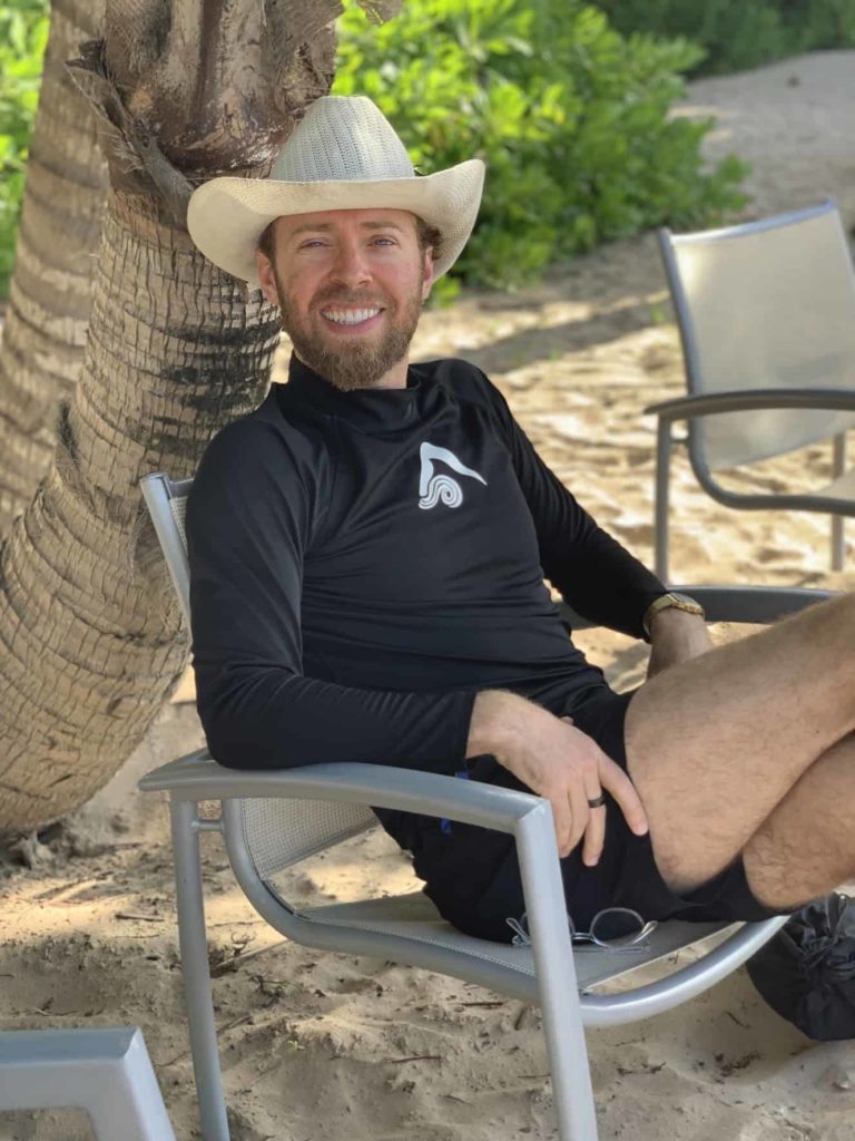 White guy (me) wearing a cowboy hat, black long-sleeve shirt, and shorts sitting in a chair on the St Regis Bahia Beach Resort in the Caribbean