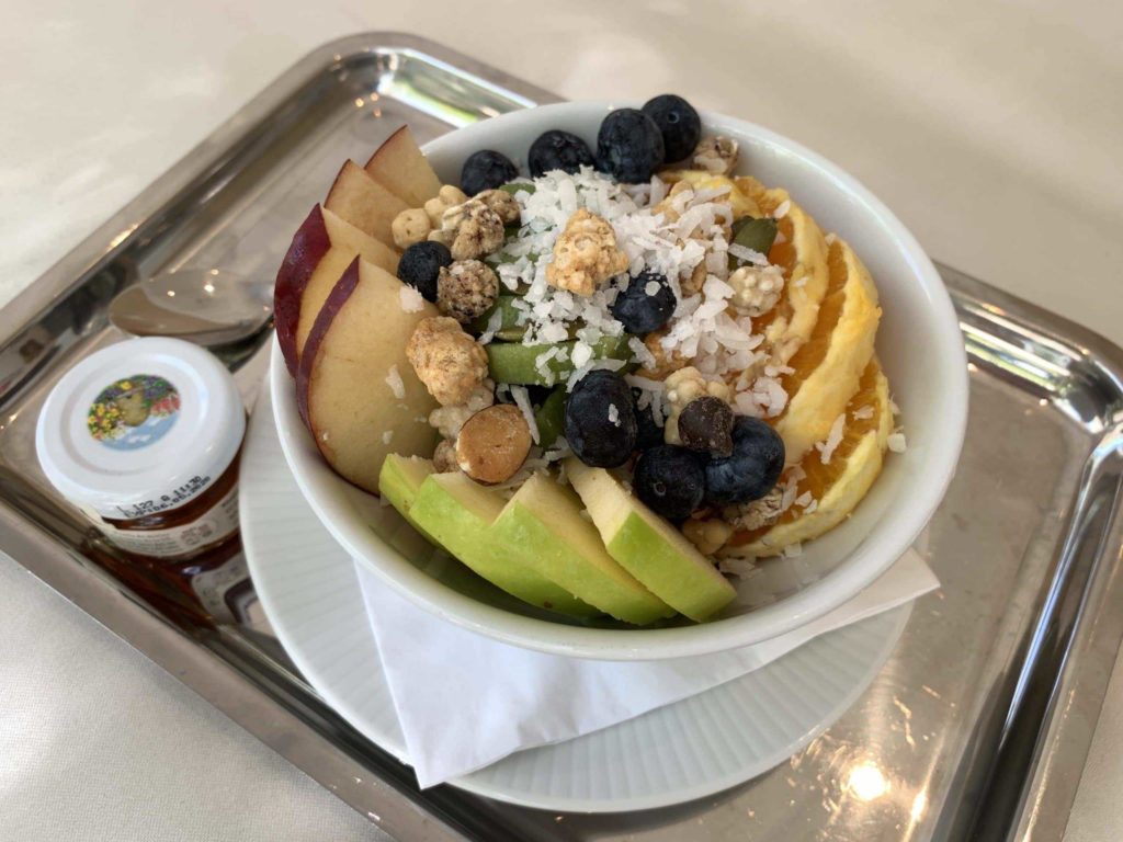 White bowl with apple, pear, blueberries, some nuts, and oranges inside on a silver tray