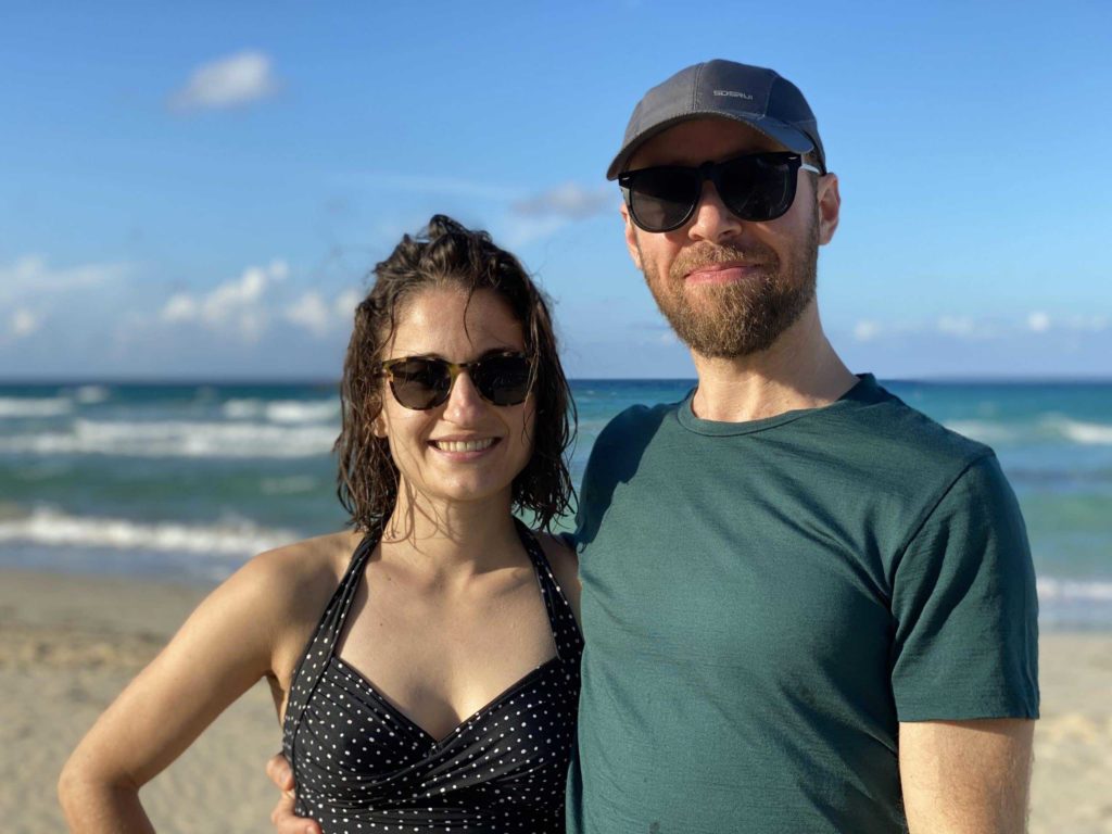 Two people smiling and wearing sunglasses with ocean / beach behind them