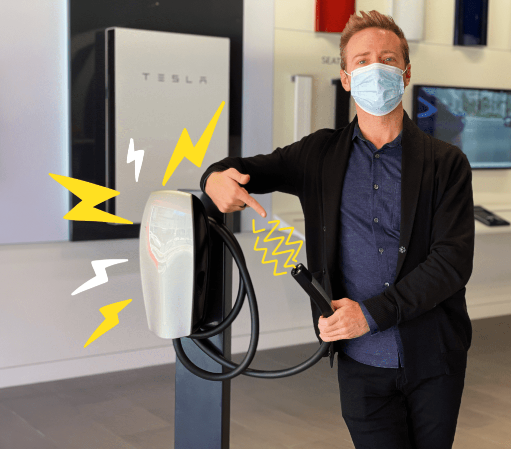 Man wearing a mask inside a Tesla dealership, leaning on a Tesla charging stand and holding the electric car charging plug