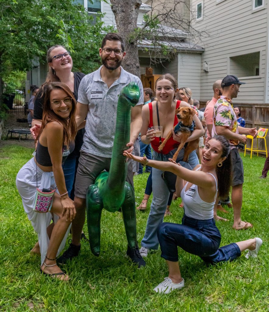 group of adults at a dinosaur party and one man in the middle is riding an inflatable brontosaurus