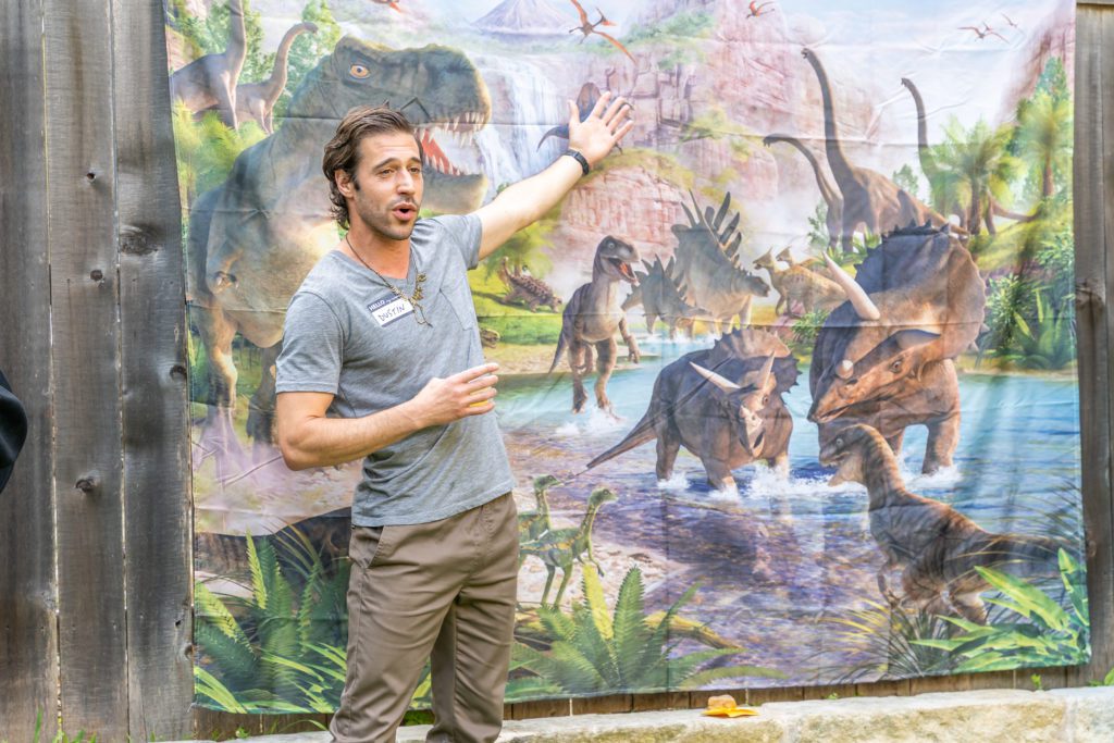 Dinosaur Whisperer aka Dustin Growick standing in front of a dinosaur tapestry (man wearing a gray shirt and brown pants)