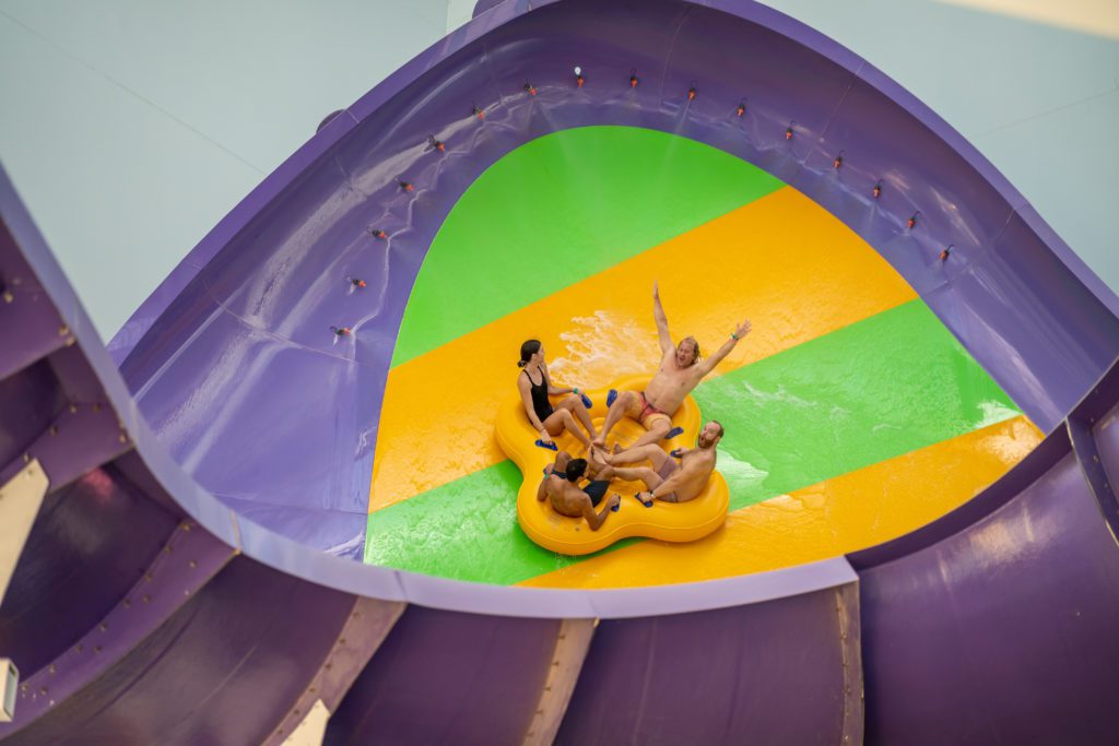 four adults inside a large yellow rube riding a water slide