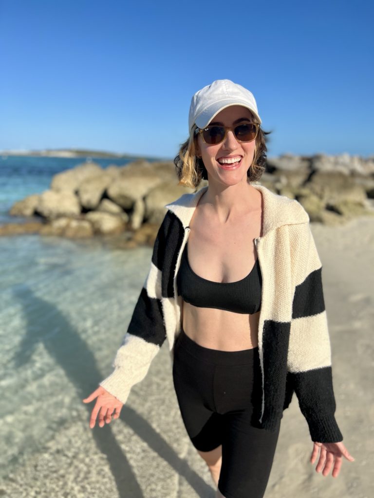 woman wearing a sweater and bathing suit walking on the white sand beach with nice blue water and rocks behind her
