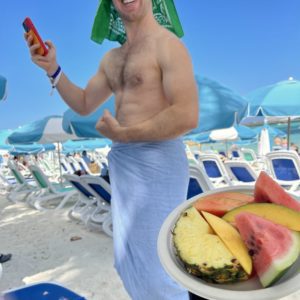 topless man holding a phone and a plate of fruit with watermelon and pineapple and mango
