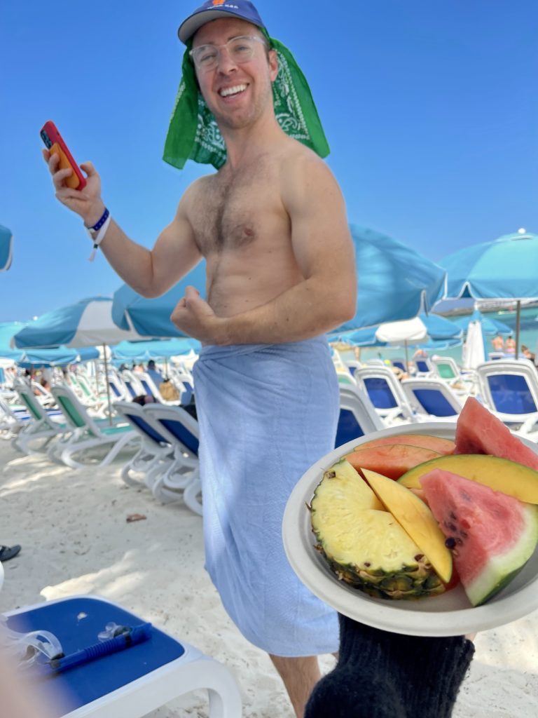 topless man holding a phone and a plate of fruit with watermelon and pineapple and mango