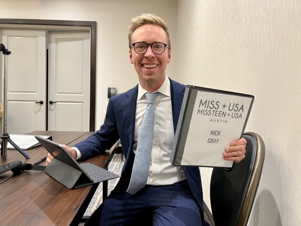 blonde man in glasses and a suit holding a binder that says NICK GRAY MISS AUSTIN